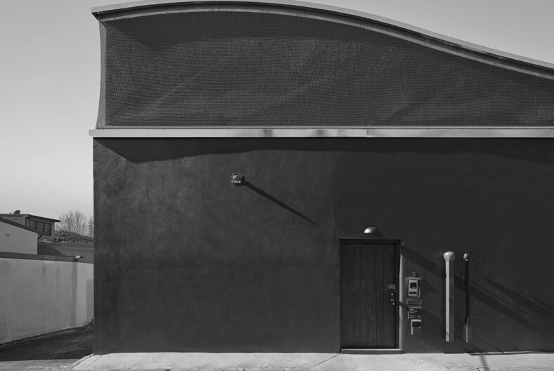 Black-and-white exterior wall with a small door, utility boxes, and long shadows across the facade.