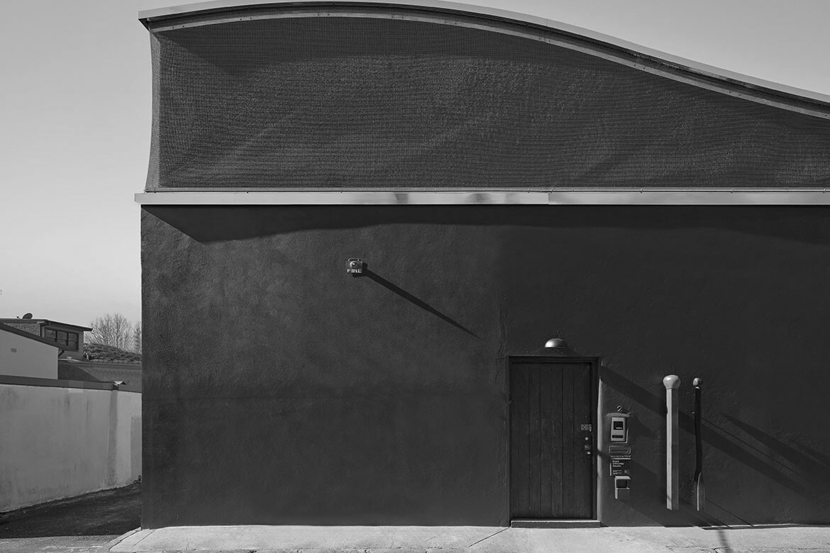 Black-and-white exterior wall with a small door, utility boxes, and long shadows across the facade.