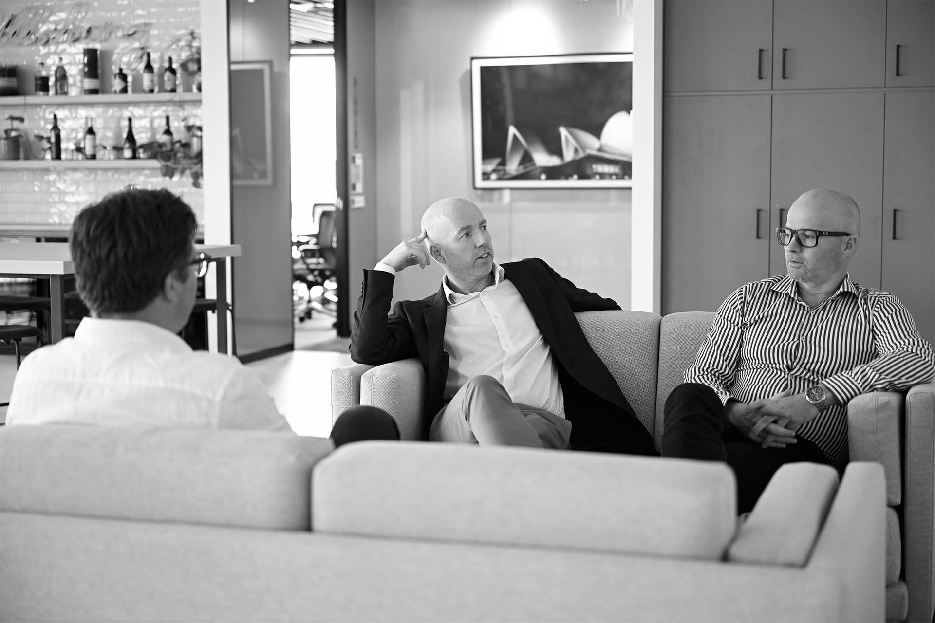 meet-the-team-wsce - WSce Three men in business attire sit on a sofa in a modern office lounge, engaged in a discussion.